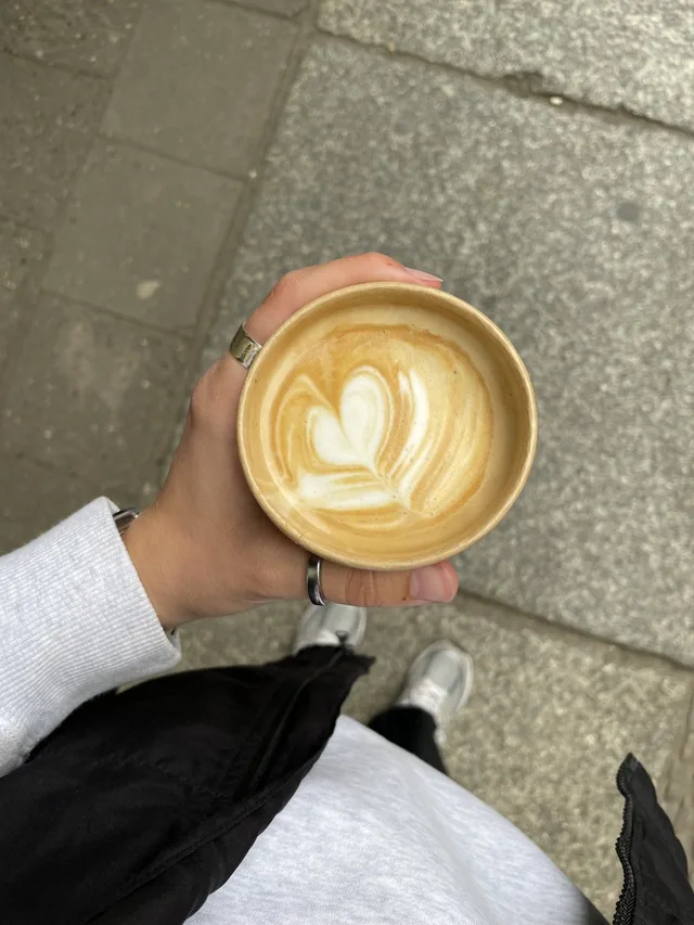 A hand holding a takeaway latte with a leaf pattern in the milk foam